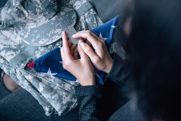 cropped view of woman on couch with military clothing, tag dog and foded american flag at home