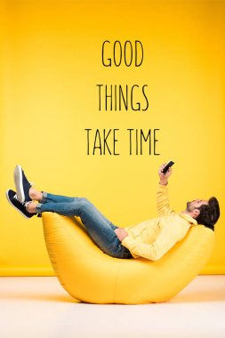 man relaxing on bean bag chair and using smartphone on yellow background with good things take time inspiration clipart