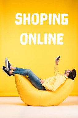 Fasulye koltukta dinlenen ve sarı arka planda akıllı telefon kullanan bir adam online alışveriş yapıyor.