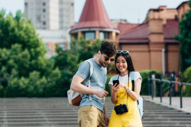 smiling and handsome man and asian woman looking at smartphone clipart