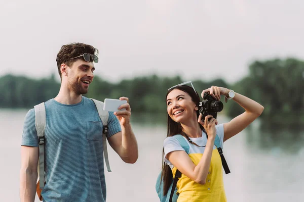 Uśmiechnięta Asian Kobieta Trzyma Aparat Cyfrowy Przystojny Mężczyzna Trzyma Smartphone — Zdjęcie stockowe