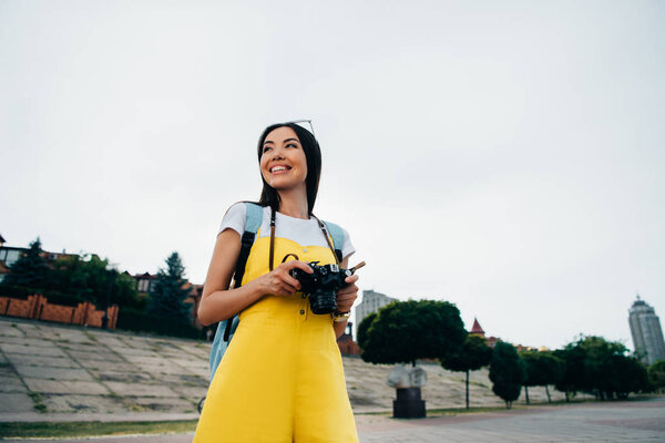 smiling and asian woman holding digital camera and looking away 