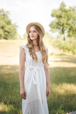 hasır şapka ve beyaz elbise ile güzel kız çayır üzerinde duran ve uzağa bakıyor
