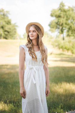 beautiful girl with straw hat and white dress standing on meadow and looking away clipart