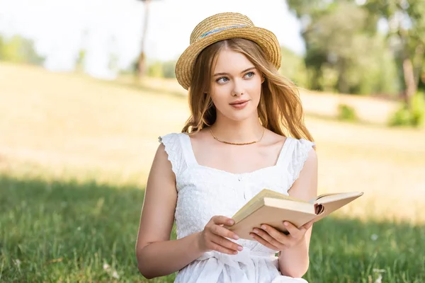 Όμορφο Κορίτσι Λευκό Φόρεμα Και Ψάθινο Καπέλο Κρατώντας Βιβλίο Ενώ — Φωτογραφία Αρχείου