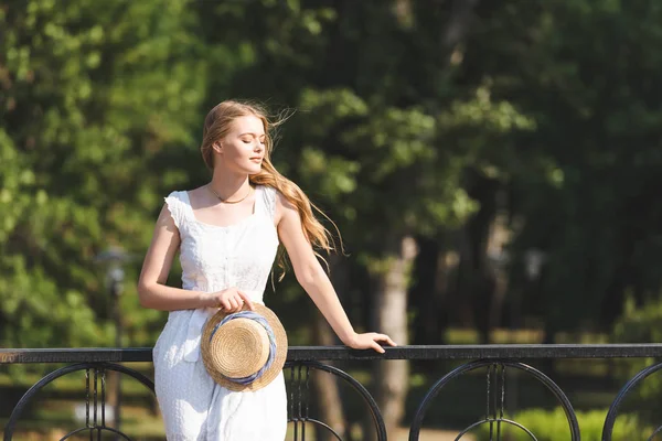 Hermosa Chica Vestido Blanco Sosteniendo Sombrero Paja Mientras Apoya Pasamanos — Foto de Stock