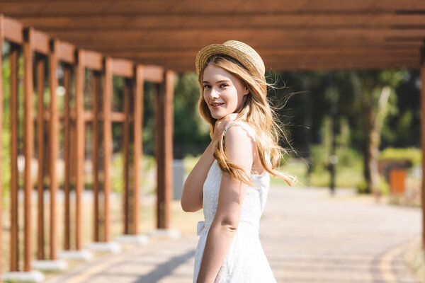 selective focus of beautiful girl in white dress and straw hat walking near wooden construction and looking at camera