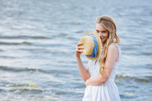 beautiful girl holding straw hat near face, smiling and looking at camera