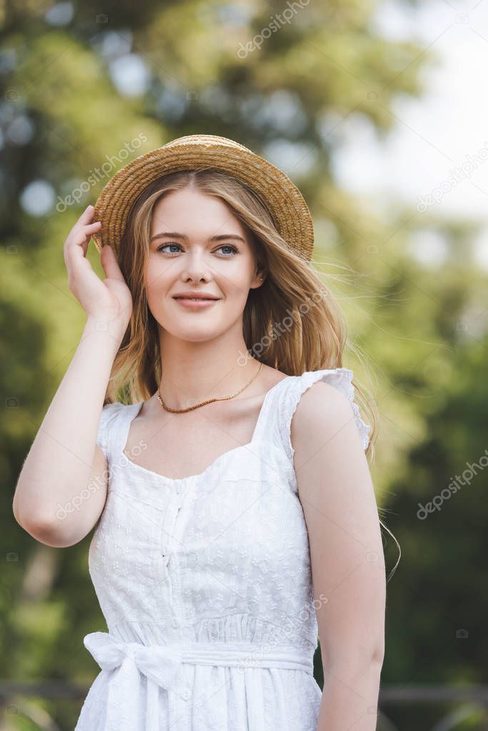 front view of beautiful young girl in white dress touching straw hat while smiling and looking away