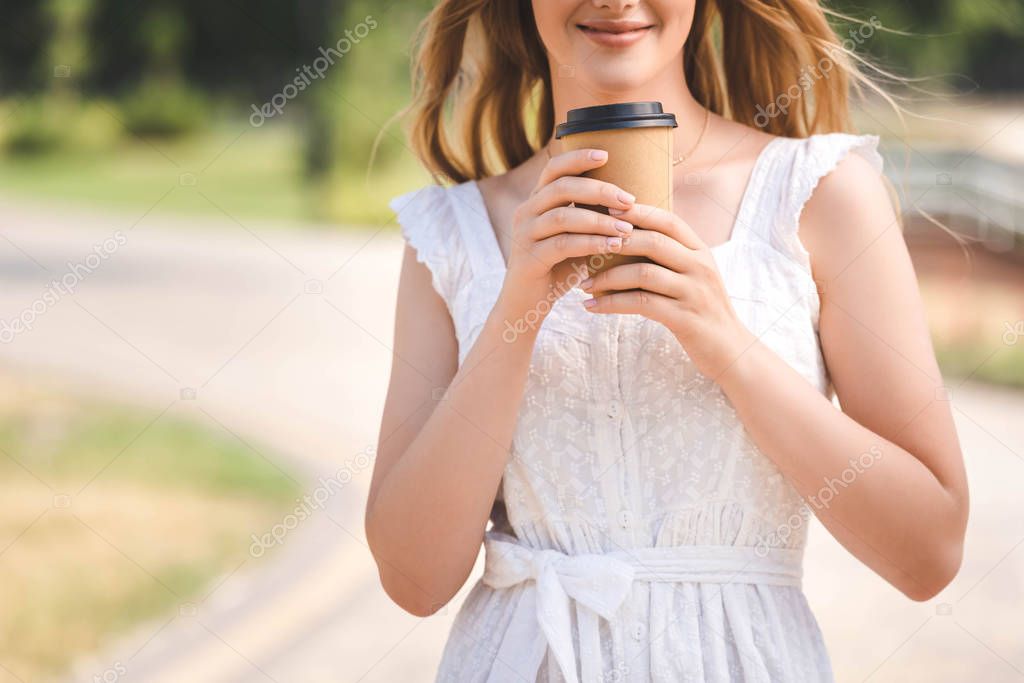 cropped view of young girl in white dress and straw hat smiling and holding paper coffee cup 