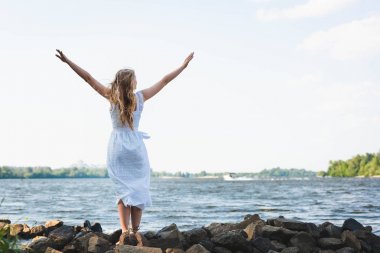 full length view of girl in white dress standing on rocky river shore with hands in air clipart