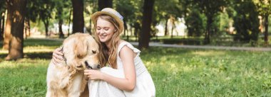 çayırda otururken beyaz elbise ve hasır şapka petting altın retriever güzel kız panoramik çekim