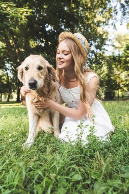 beyaz elbise ve hasır şapka güzel genç kız gülümseyerek, çayırda oturan ve köpek bakarken altın retriever petting