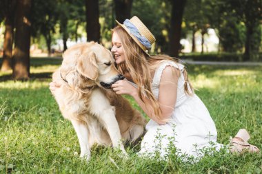 beyaz elbise ve hasır şapka ile güzel genç kız gülümseyerek ve kapalı gözlerle çayırda otururken altın retriever besleme 