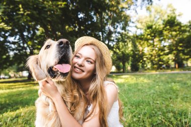 beyaz elbise ve hasır şapka güzel kız çayırda otururken, gülümseyerek ve köpek bakarken altın retriever sarılma