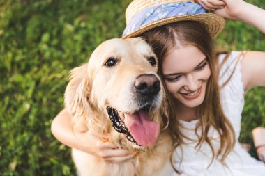 beyaz elbise ve hasır şapka ile güzel genç kız yüksek açı görünümü çayırda otururken, gülümseyerek ve uzağa bakarken altın retriever sarılma