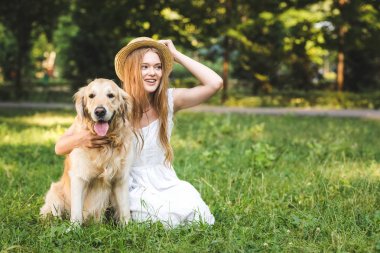 beyaz elbiseli güzel genç kız, gülümseyerek, çayırda otururken ve uzağa bakarken altın av köpeğine sarılırken hasır şapkaya dokunuyor