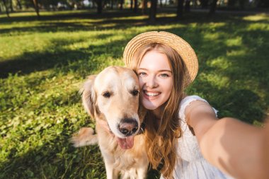 beyaz elbise ve hasır şapka güzel genç kız altın retriever sarılma ve çayırda otururken selfie alarak, gülümseyerek ve kameraya bakarak