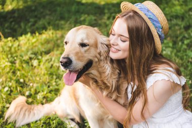 beyaz elbise ve hasır şapka güzel kız çayırda otururken ve köpek bakarken altın retriever sarılma