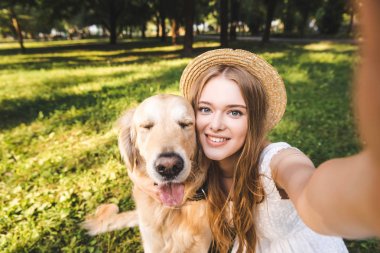 beyaz elbiseli ve hasır şapkalı genç kız altın av köpeğine sarılıyor ve çayırda otururken selfie çekiyor, gülümsüyor ve kameraya bakıyor