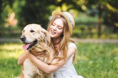 selective focus of beautiful girl in white dress and straw hat hugging golden retriever and smiling while sitting on meadow clipart