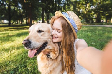 beyaz elbiseli ve hasır şapkalı genç kız altın av köpeğine sarılıyor ve çayırda otururken ve kapalı gözlerle gülümserken selfie çekiyor