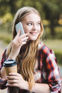 kağıt kahve fincanı tutan ve akıllı telefon konuşurken gülümseyen ve uzağa bakarak rahat giysiler içinde güzel kız portre çekimi