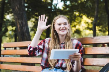 gündelik giysiler içinde güzel kız parkta ahşap bankta oturan, dijital tablet kullanarak ve el sallayarak