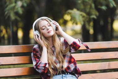 beautiful girl in casual clothes listening to musing on headphones and looking away while sitting on wooden bench in park