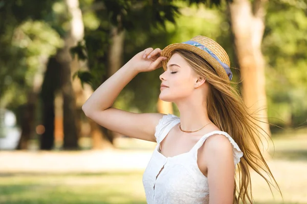 美丽的年轻女孩在白色礼服触摸草帽 而站在公园与闭着眼睛 — 图库照片