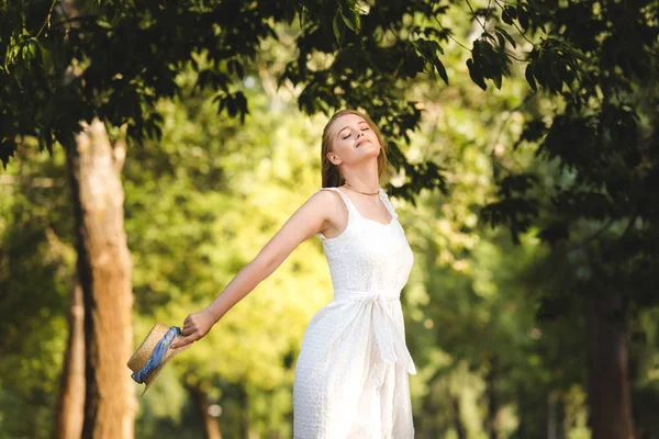 Hermosa Chica Vestido Blanco Sosteniendo Sombrero Paja Mientras Sonríe Pie — Foto de Stock