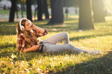 güneş ışığında çayırda yatan ve kulaklıkmüzik dinlerken rahat giysiler içinde güzel kız tam uzunlukta görünümü