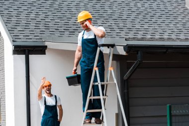 neşeli handyman el sallayarak seçici odak ve akıllı telefon konuşurken iş arkadaşı bakarak 