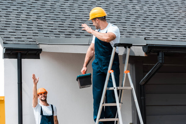 selective focus of workman waving hand and looking at coworker standing on ladder with toolbox 