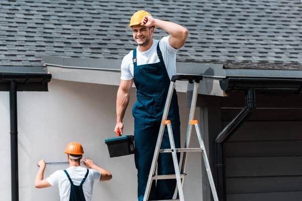 selective focus of happy repairman touching helmet and standing with toolbox on ladder near coworker 