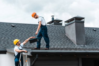 çatıda duran yakışıklı meslektaşına alet çantası veren kask handyman 