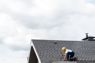 repairman in helmet holding hammer while repairing roof  clipart