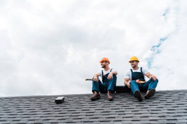 cheerful handymen in helmets sitting on rooftop  clipart