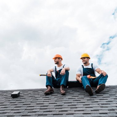 cheerful handymen in helmets and uniform sitting on rooftop  clipart