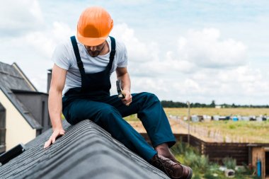 turuncu kask içinde handyman seçici odak çatıda oturan ve çekiç tutarak 