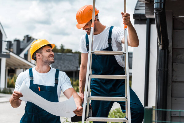 happy builder looking at coworker in helmet standing on ladder 