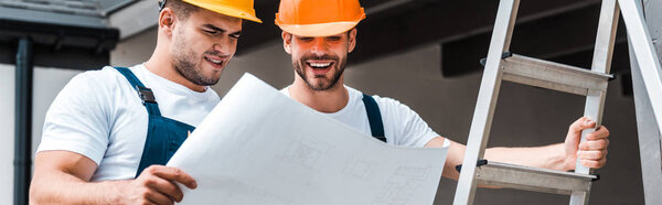 panoramic shot of happy builders in helmets looking at paper 