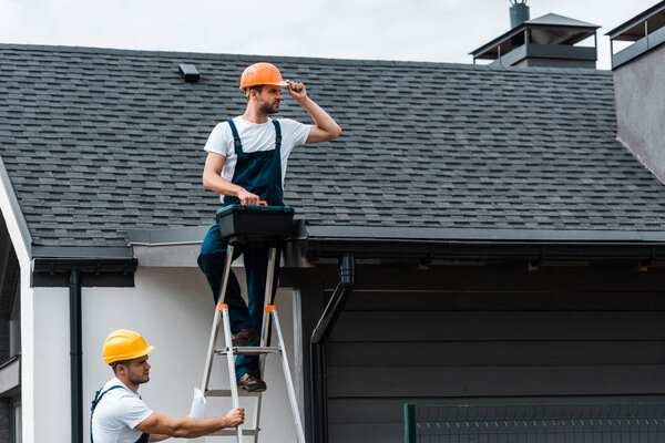 handsome repairman sitting on roof and holding toolbox near coworker in helmet 