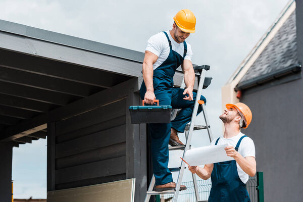 cheerful architect pointing with finger at paper near happy coworker in helmet 