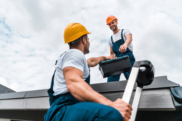 low angle view of handyman in helmet giving toolbox to happy colleague 