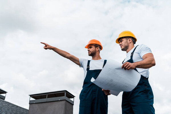 low angle view of handsome architect pointing with finger near coworker in helmet holding paper 
