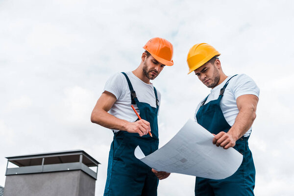 low angle view of handsome builders in uniform and helmets looking at paper with plan 