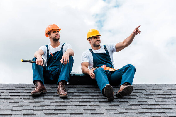 happy handyman pointing with finger while sitting on rooftop with coworker 