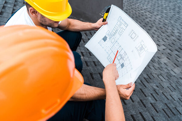selective focus of architect in helmet holding pencil near blueprint and coworker 