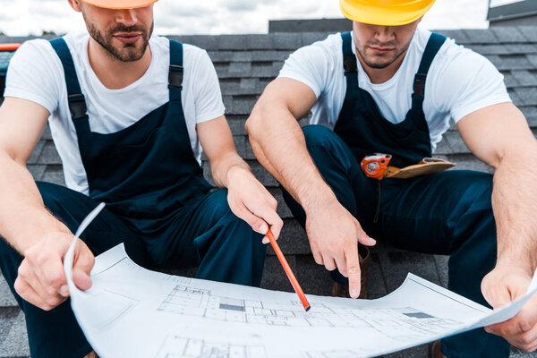 cropped view of architect in helmet holding pencil near blueprint and coworker pointing with finger 